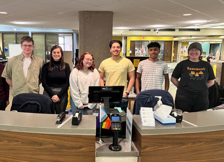 Six college-aged student assistants smiling for a photo standing behind the Library Service Desk.